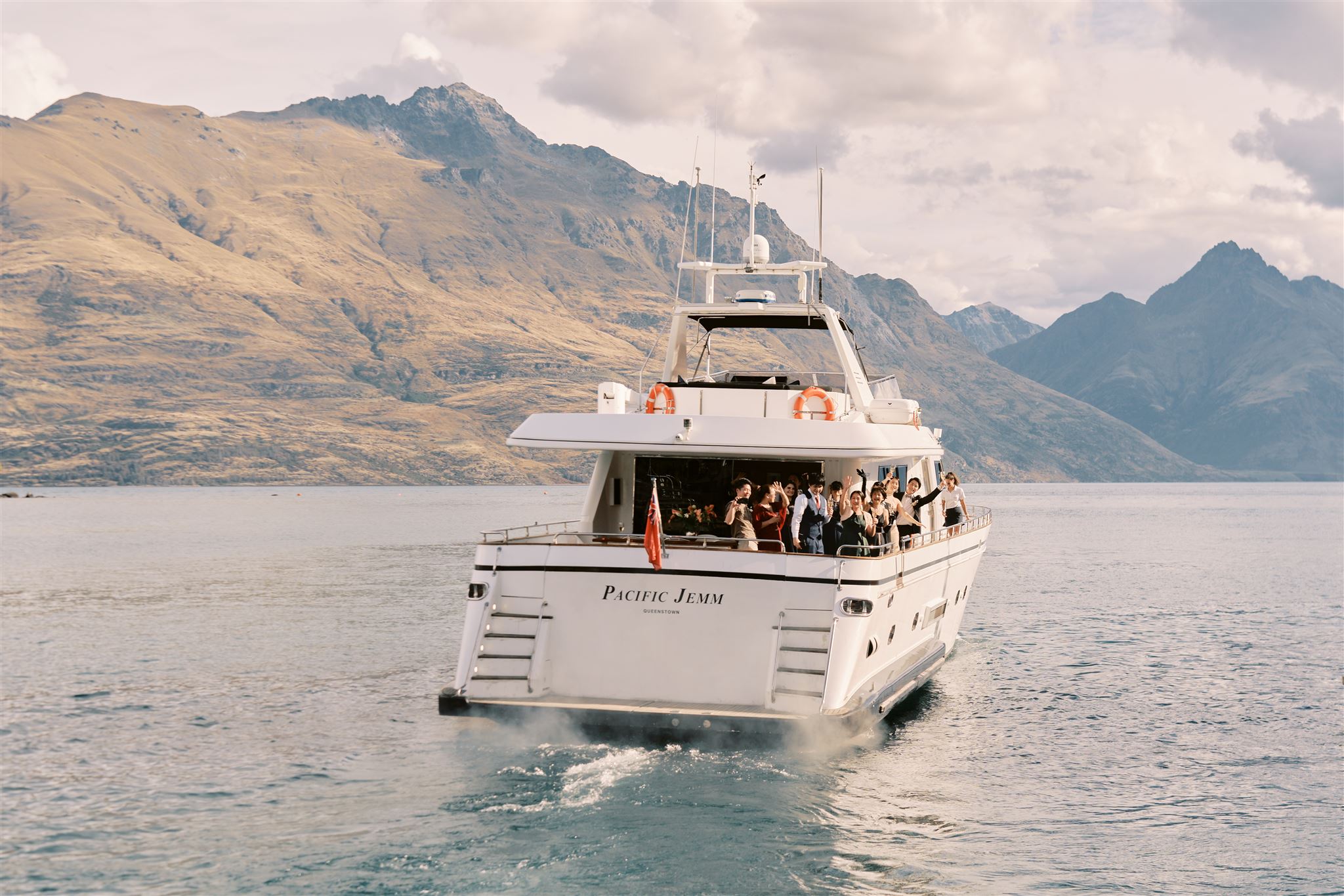 Queenstown New Zealand Heli Wedding Elopement Photographer クイーンズタウン　ニュージーランド　エロープメント 結婚式 | A group of passengers enjoying a boat tour on a cloudy day with majestic mountains in the background, captured by Yuri, the Queenstown Wedding Photographer.